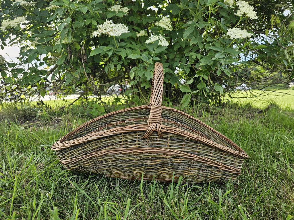 Bootsförmiger Blumenkorb aus Weide mit Bügelhenkel in der Mitte im Gras vor einem blühenden Holunderbusch