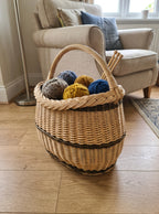 Medium oval shopping basket with handle, handwoven from willow with a braided rim, holding balls of yarn and knitting needles beside an armchair.