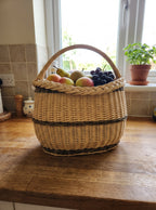 Handwoven willow market basket with a braided rim holding apples, pears and grapes on a kitchen counter in front of a window.