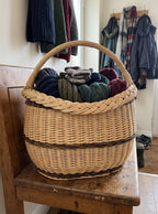 Oval willow basket with a braided rim filled with knitted hats and gloves on a bench in a hallway under hanging coats.