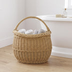 Medium handwoven wicker basket with a handle holding rolled white towels on a wooden floor beside a freestanding bath.