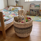 Wicker harvest basket with a tall handle used as a toy basket for teddy bears and wooden blocks beside a child's bed
