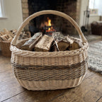 Large wicker kindling basket with handle filled with split firewood in front of a lit fireplace in a rustic living room