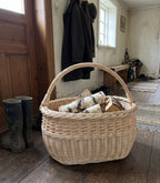 Oval willow harvest basket with handle holding birch logs in a farmhouse entryway next to rubber boots and a coat rack