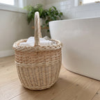 Handwoven wicker basket with a tall handle holding folded towels on a wooden floor beside a freestanding bathtub