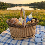 Large oval wicker picnic basket with handle packed with bread, cheese, apples and a bottle on a checked blanket by a lake at sunset
