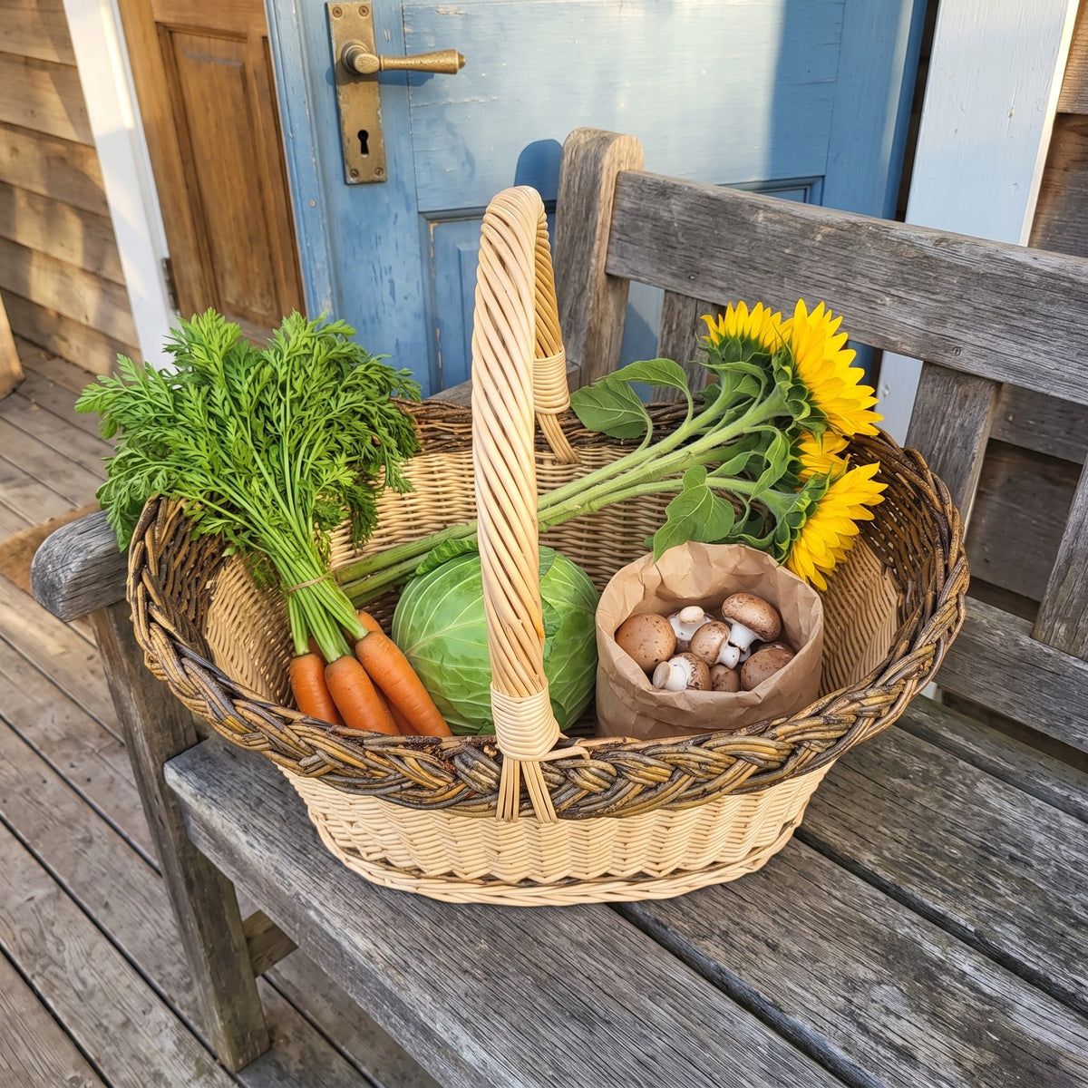Handwoven oval picnic basket with a tall handle full of carrots, cabbage, mushrooms and sunflowers on a wooden garden bench