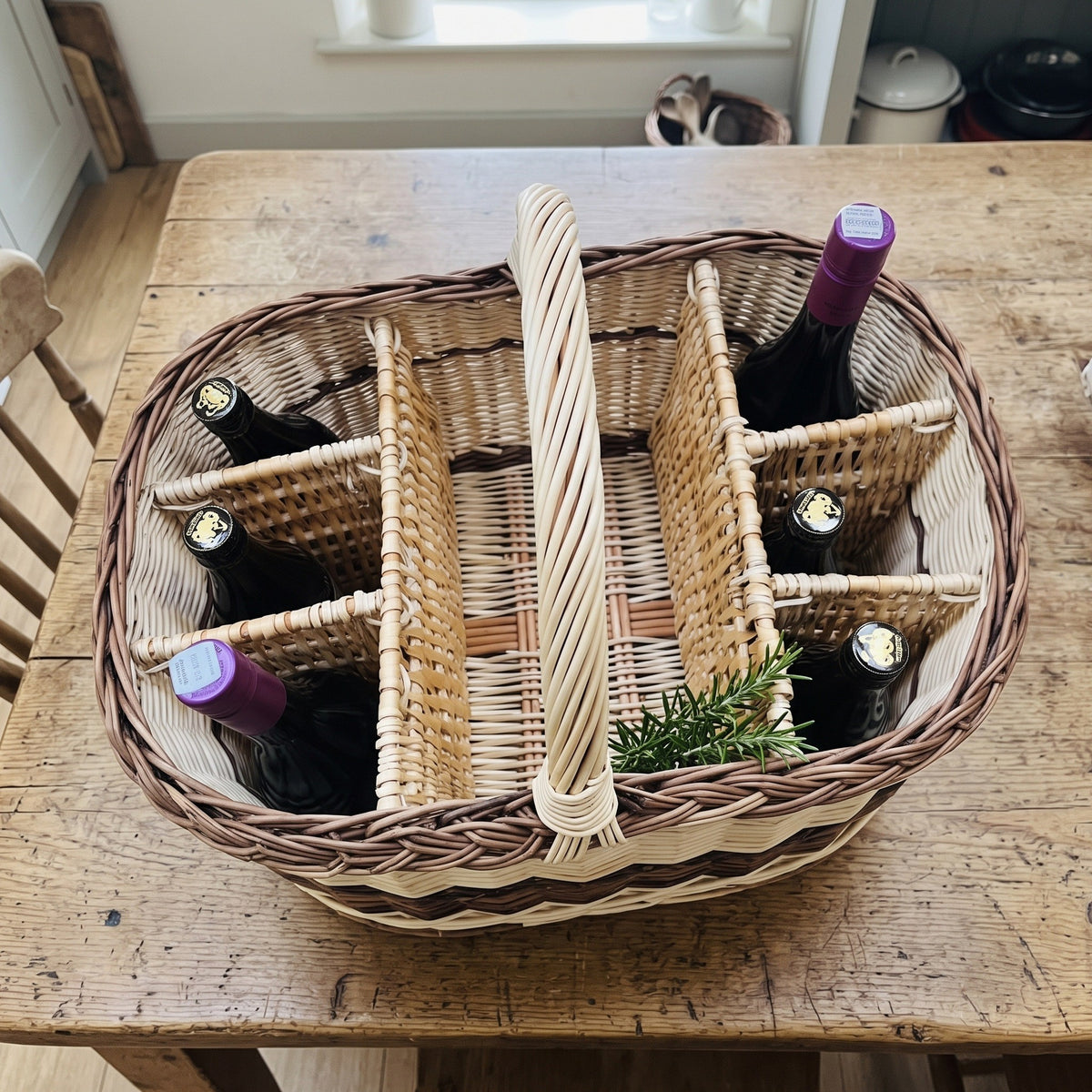 Top view of the handwoven willow wine carrier with dividers on a farmhouse table, six wine bottles in separate compartments and a rosemary sprig.