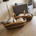 Pair of handwoven willow storage baskets with handles holding cushions and wool throws on a wooden floor beside a grey sofa in a living room