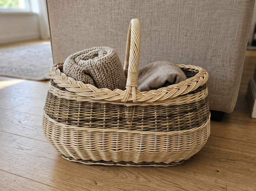 Large oval willow basket with handle holding folded knitted blankets on a wooden floor beside a sofa