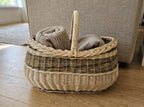Large oval willow basket with handle holding folded knitted blankets on a wooden floor beside a sofa