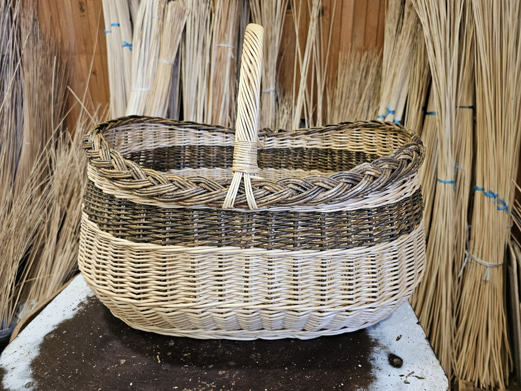 Large oval willow shopping basket with tall handle on a workbench in front of bundles of willow rods in the workshop