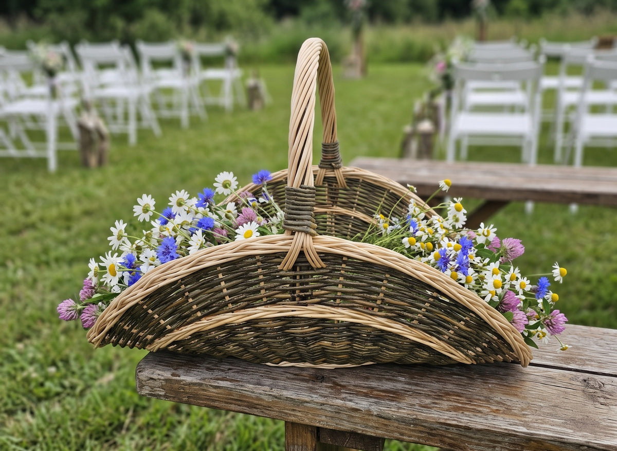 Unpeeled willow garden trug with a tall handle filled with wildflowers on a wooden bench at an outdoor rustic wedding ceremony with white chairs.