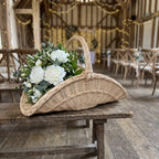 Boat-shaped handwoven willow flower basket holding white peonies and greenery on a rustic bench at a barn wedding venue with fairy lights.