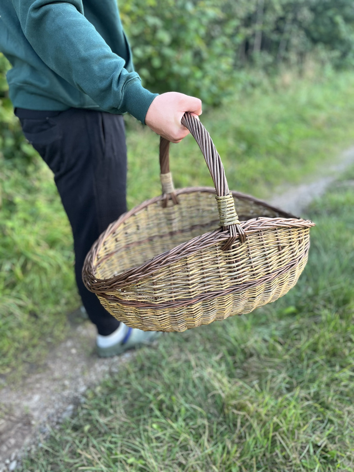 The willow basket carried in one hand by a person walking along the grass verge, showing its size in use.