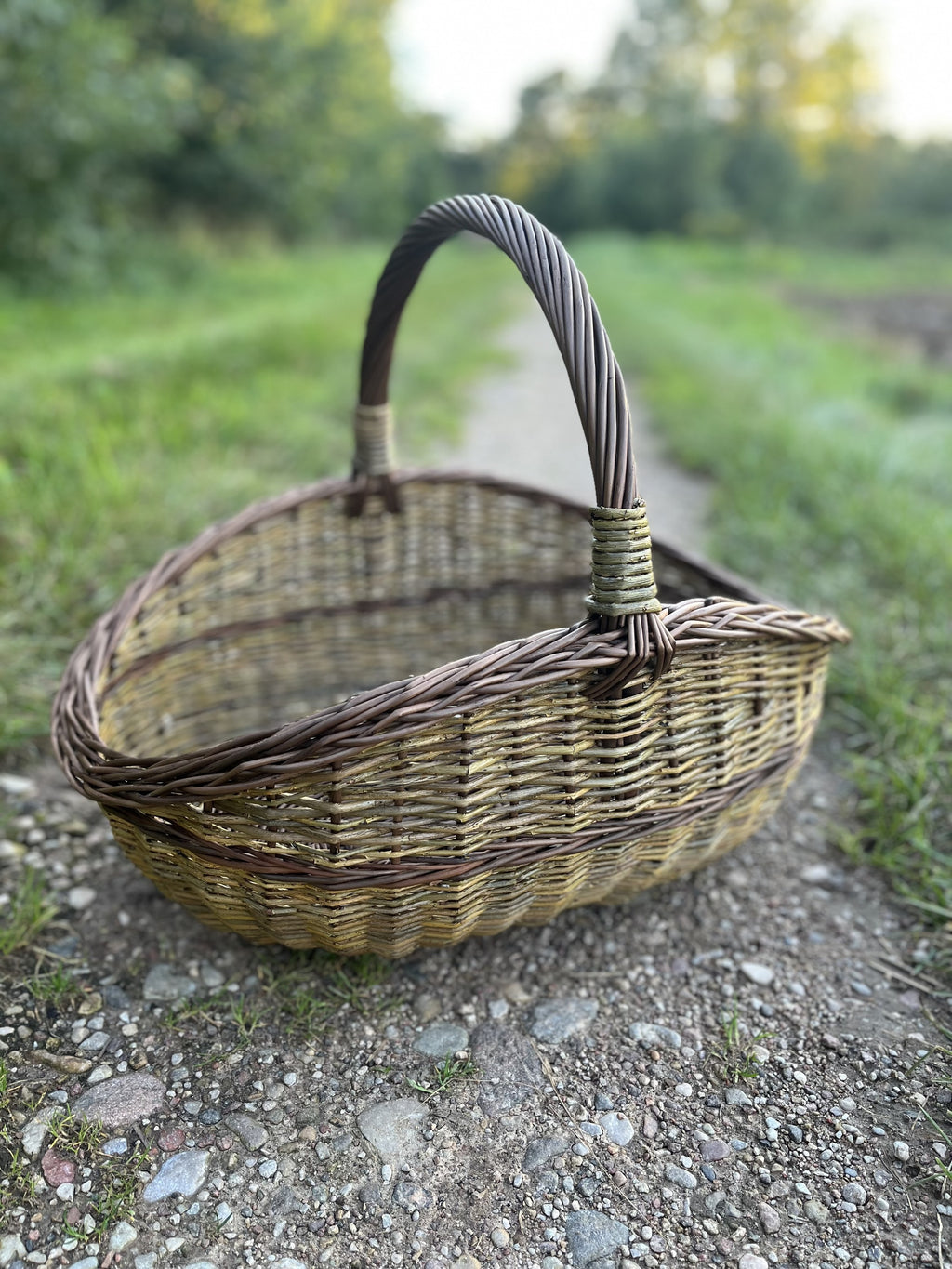 Large handwoven unpeeled willow gathering basket with a twisted handle, standing on a gravel country track beside the grass.