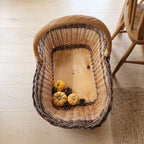 Top view of the oversized wicker harvest basket showing its solid wooden base with small pumpkins and pine cones beside a wooden chair.