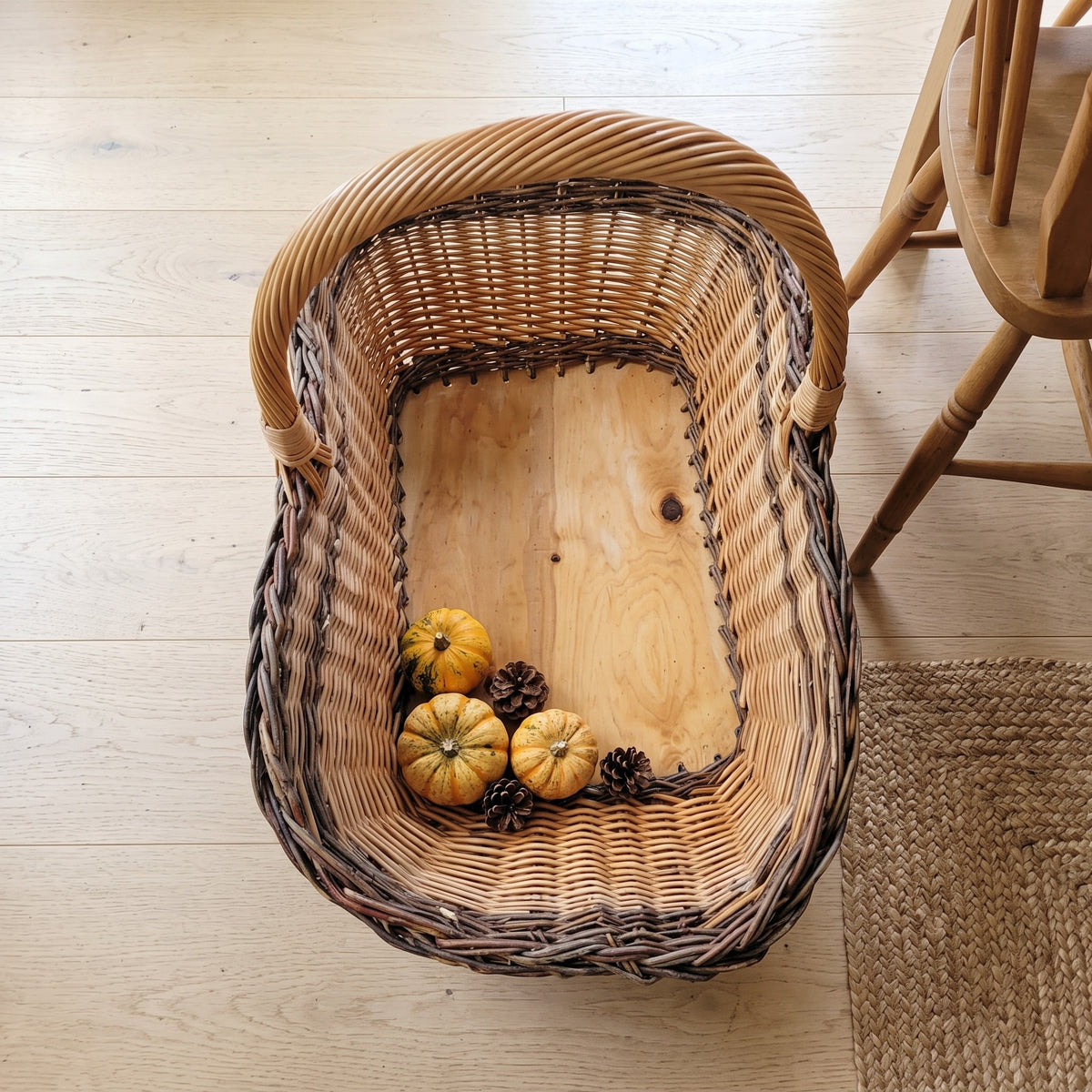Top view of the oversized wicker harvest basket showing its solid wooden base with small pumpkins and pine cones beside a wooden chair.