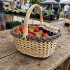 Wicker basket with handle at a farmers market stall, holding fresh strawberries and a jar of homemade jam with a linen-covered lid.