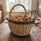 Handwoven bread basket with handle and braided rim filled with croissants and rolls on a linen cloth at a sunny breakfast table.