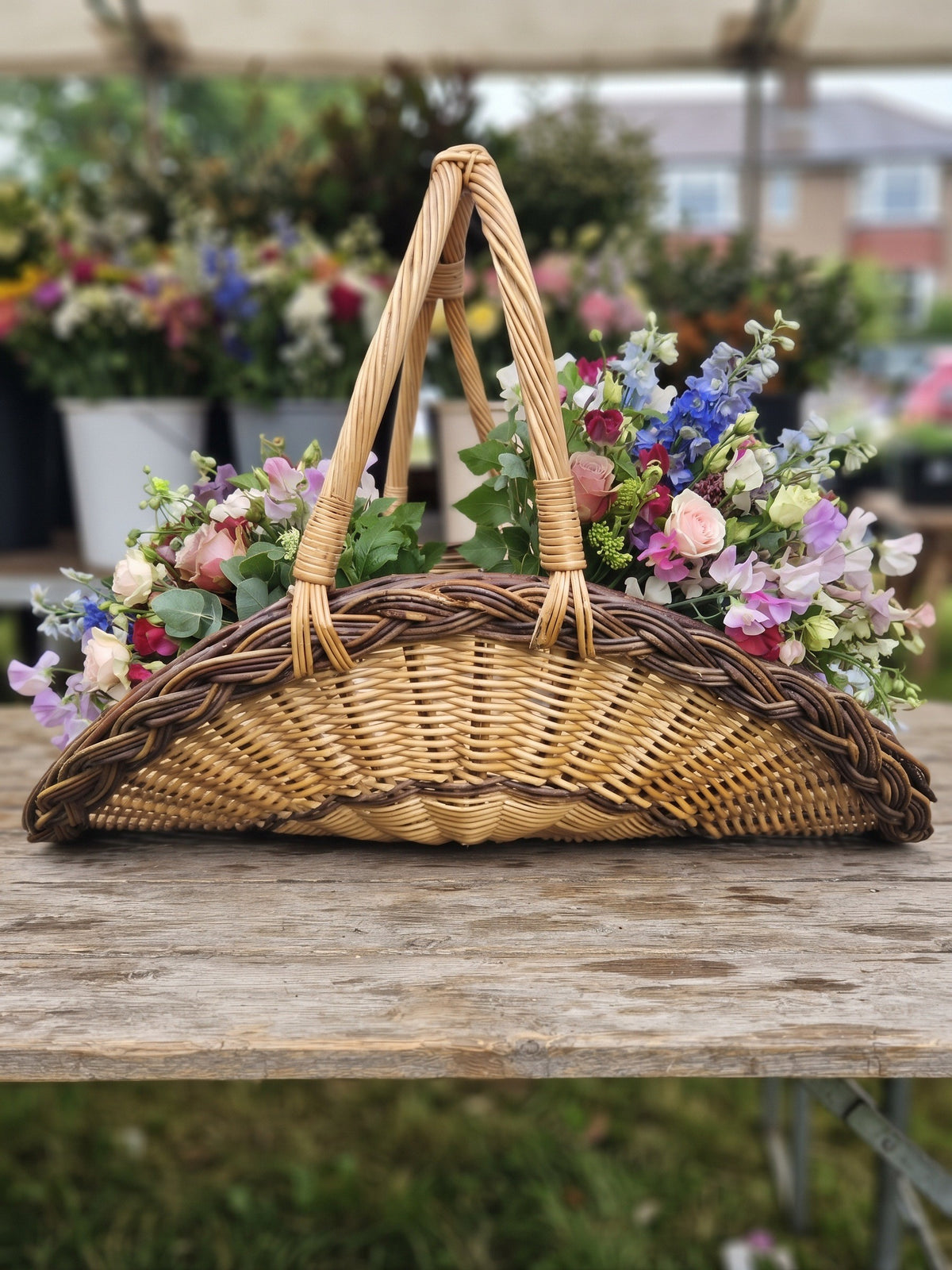 Wicker flower trug with a tall double arched handle, handwoven from willow and filled with mixed garden flowers on a rustic table at a flower market.