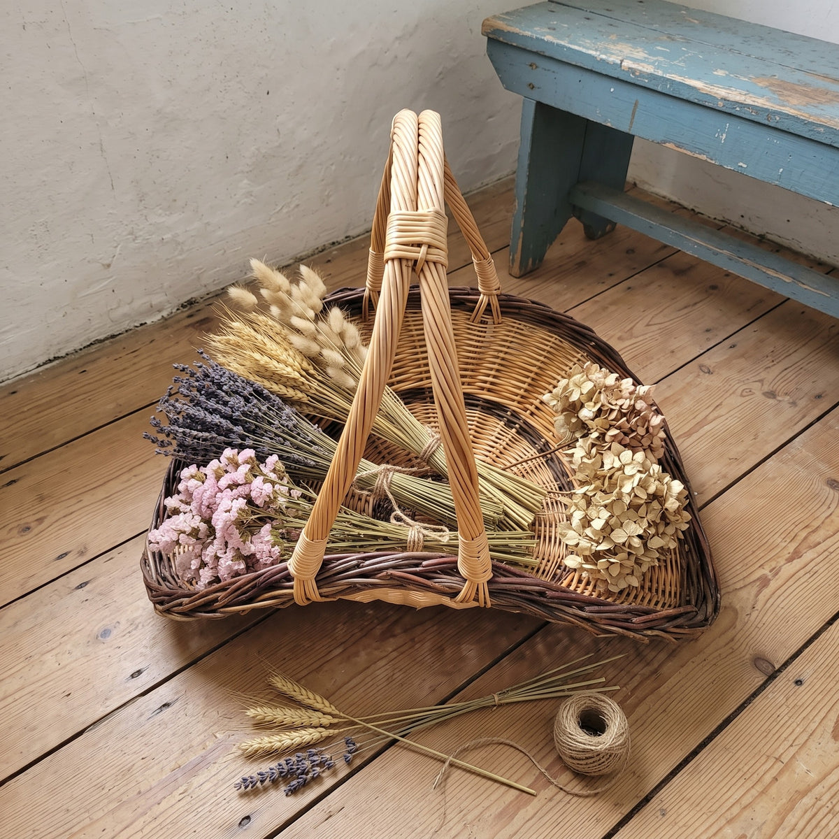 Handwoven willow trug on small wooden feet filled with dried lavender, wheat and hydrangeas on a wooden floor beside a bench and twine.