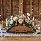 Wicker table centerpiece on wooden feet with a tall arched handle, filled with roses and greenery on a candlelit wedding table in a barn.