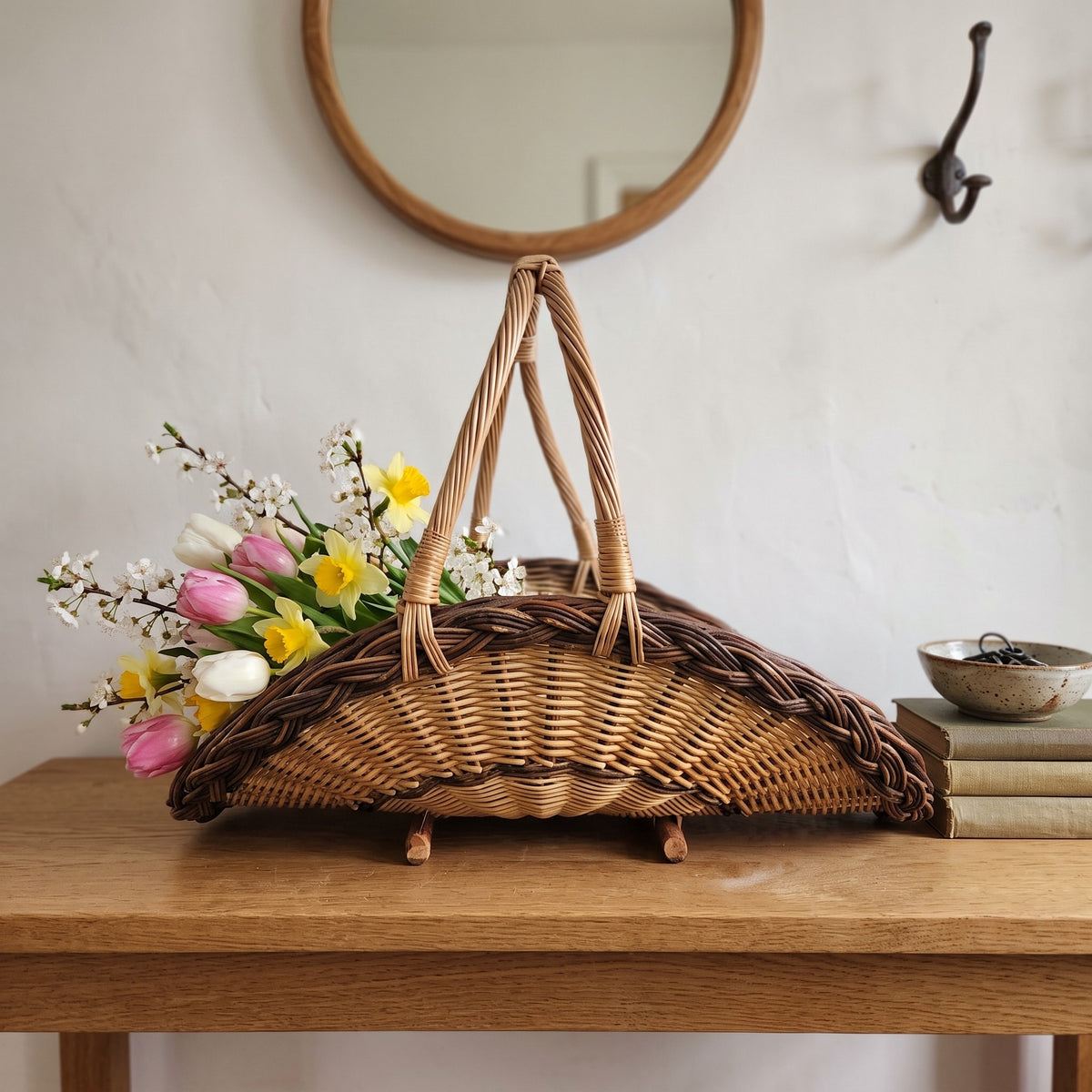 Handwoven willow flower trug on wooden feet holding tulips and daffodils on a console table in an entryway beneath a round mirror.