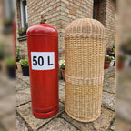 Handwoven wicker LPG sleeve standing next to an uncovered red 50 L gas cylinder in a brick courtyard, showing how the cover hides the tank.