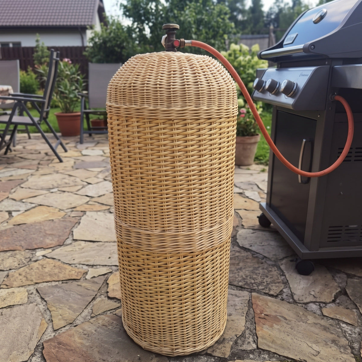 Tall wicker gas bottle sleeve on a stone patio connected to a barbecue grill, the valve and hose coming out through the opening in the top.