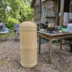 Handwoven willow LPG cylinder cover next to a camping table with a gas stove and cookware in a backyard with gravel and a wooden shed.
