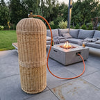 Wicker gas bottle cover beside a concrete fire pit table on a modern terrace with an outdoor sofa, the hose running to the burner.