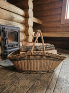 Long wicker trug with two handles holding split firewood on a plank floor beside a wood-burning stove in a log cabin