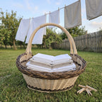 Large oval willow basket with two handles and a braided rim holding folded white linens on the grass under a washing line in an orchard