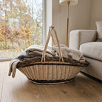 Large wicker trug with two handles used as a blanket basket, holding wool throws on a wooden floor beside a sofa in a bright living room