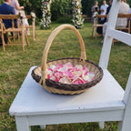 Handwoven willow flower girl basket with a tall handle filled with rose petals, set on a white chair at an outdoor garden wedding ceremony