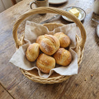 Small round wicker bread basket with a tall handle lined with linen and filled with fresh bread rolls on a wooden breakfast table