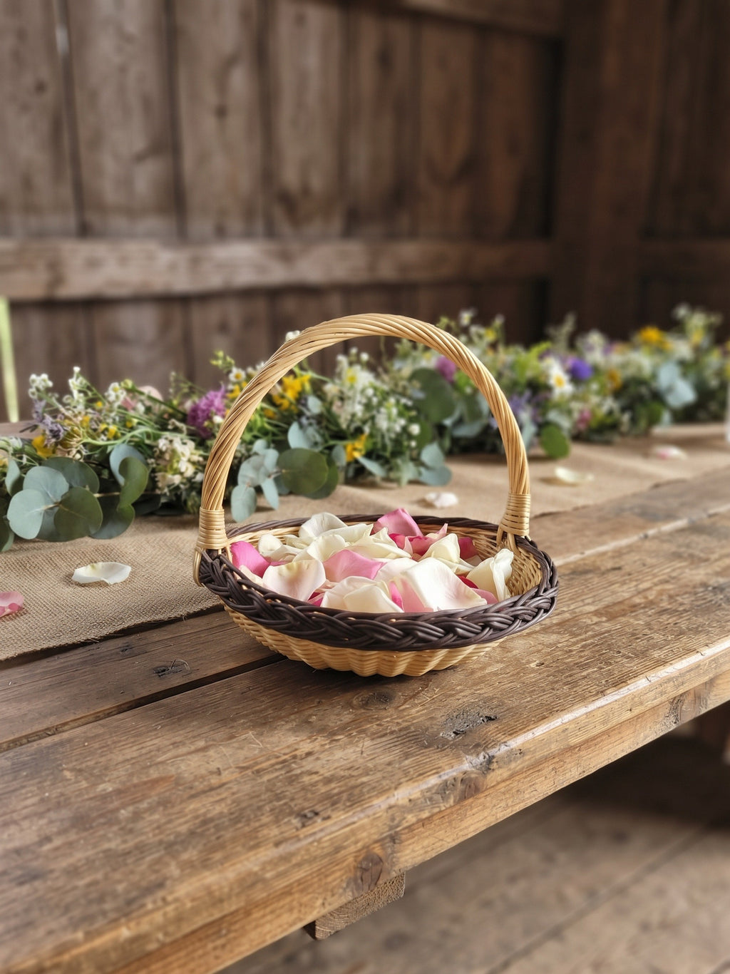 Willow flower girl basket with a tall handle filled with rose petals on a long rustic wedding table decorated with wildflowers
