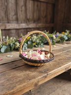 Willow flower girl basket with a tall handle filled with rose petals on a long rustic wedding table decorated with wildflowers