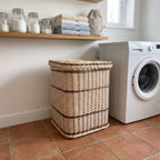 Large square wicker laundry hamper with lid standing on a terracotta tiled floor beside a washing machine in a bright laundry room