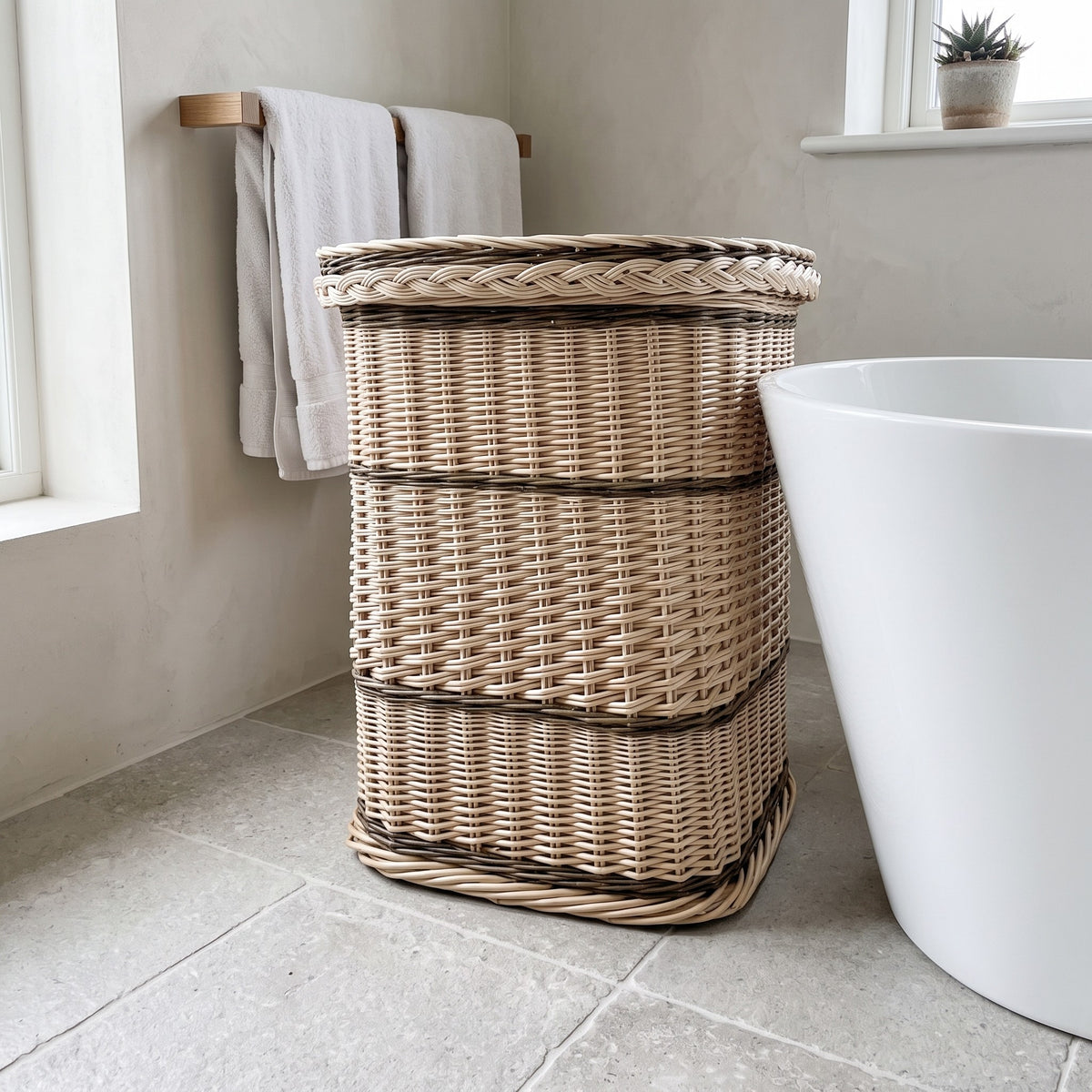 Tall handwoven willow laundry hamper with a braided lid edge next to a freestanding bathtub and towel rail in a modern bathroom