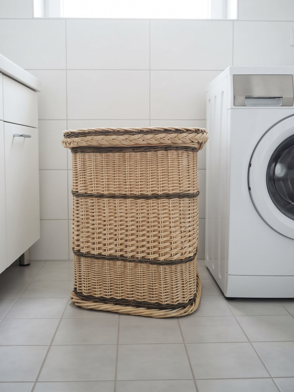 Tall handwoven laundry hamper with lid on white floor tiles between a bathroom cabinet and a washing machine