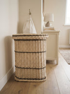Large wicker laundry hamper with lid on a wooden floor in a calm bedroom corner beside a nightstand and a hanging laundry bag