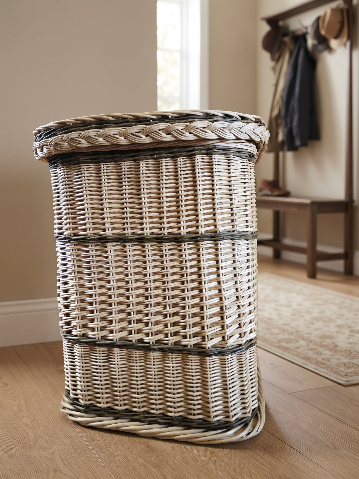 Tall wicker storage hamper with braided lid edge standing in a hallway with a coat rack and bench behind