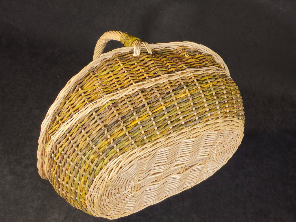 Oval willow trug basket lying on its side on a dark backdrop, showing the woven oval base and the bound handle