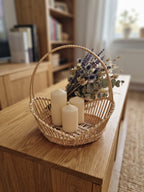 Decorative wicker basket with a tall handle holding pillar candles and a dried flower bunch on a wooden sideboard in a living room