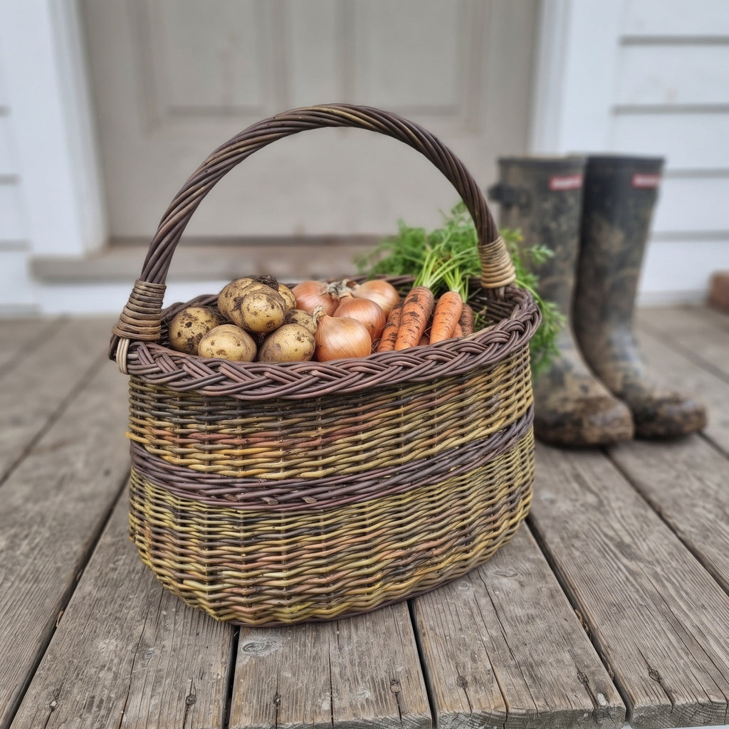 Handwoven wicker foraging basket holding potatoes, onions and carrots from the garden on a wooden porch beside muddy rubber boots.