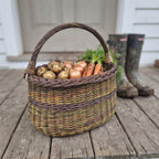 Handwoven wicker foraging basket holding potatoes, onions and carrots from the garden on a wooden porch beside muddy rubber boots.