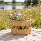 Tall willow foraging basket with an open-weave band filled with cornflowers and wild flowers on a linen cloth in a meadow by a lake.