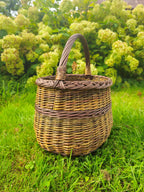 Tall handwoven willow foraging basket with a braided rim and reinforced handle standing on the grass in front of hydrangea bushes.